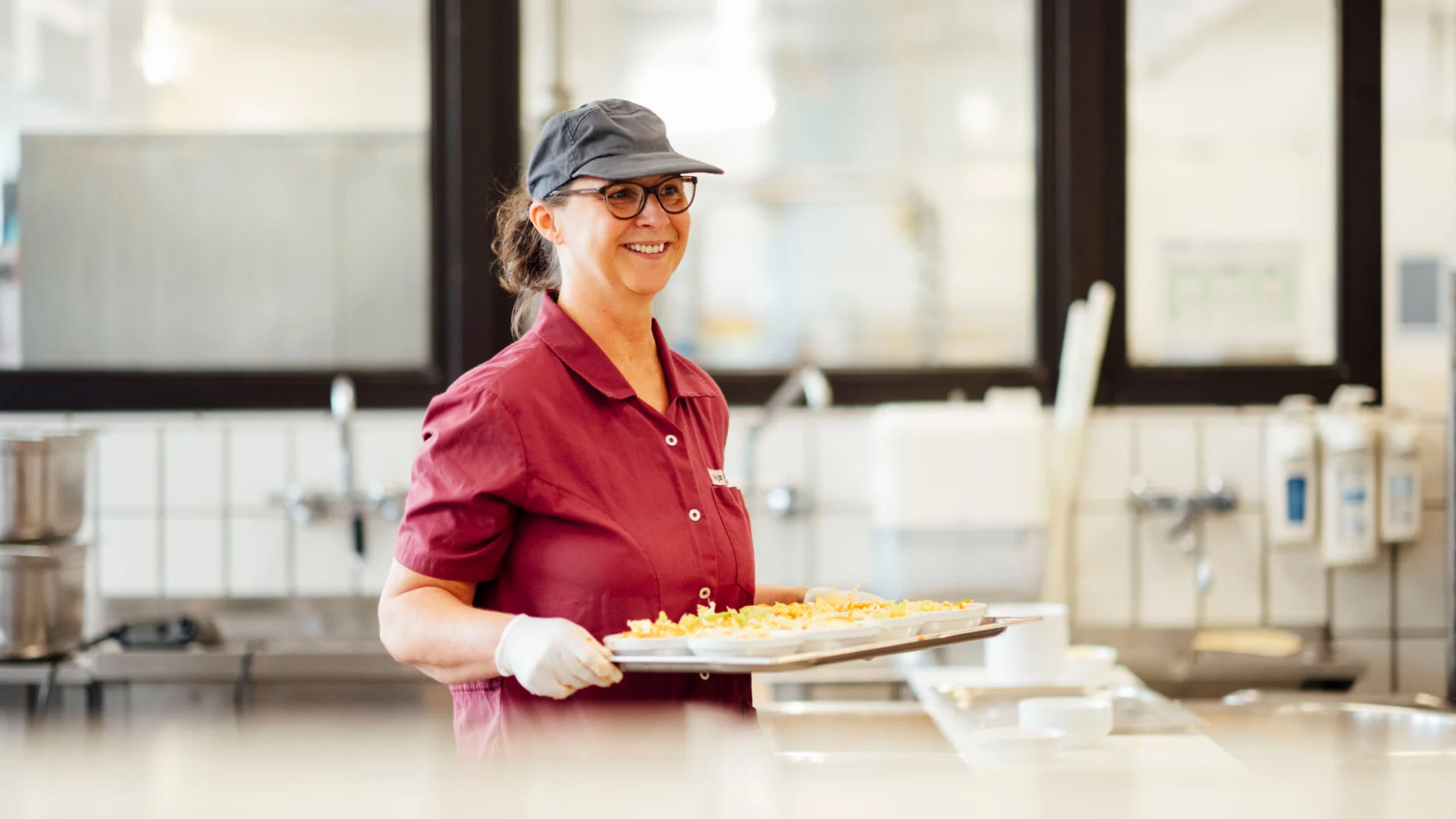 Küchenhilfe trägt Tablett mit Gerichten in der Mensaküche des Studierendenwerks Oldenburg – Küchenhilfe Job