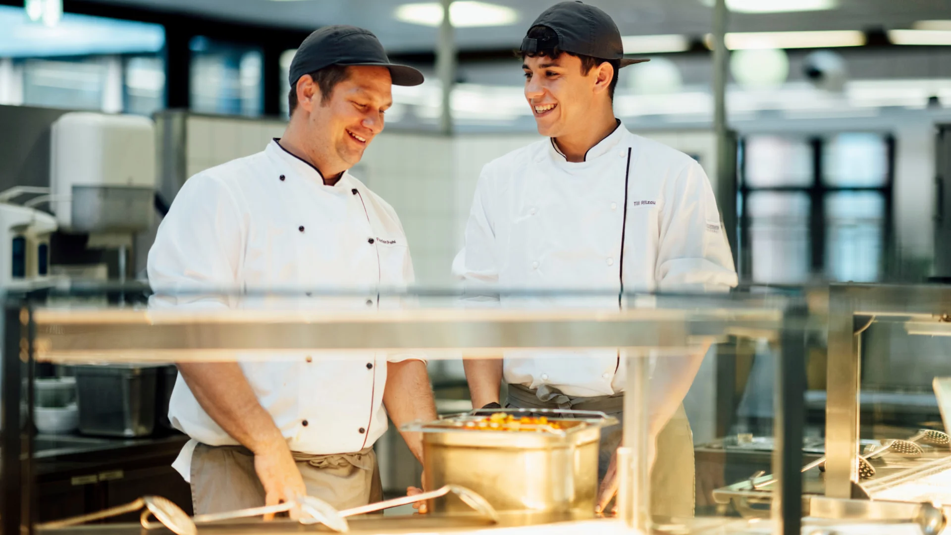 Auszubildender Koch und Küchenmeister lächeln am Ausgabecounter der Mensa im Studierendenwerk Oldenburg – Koch Ausbildung